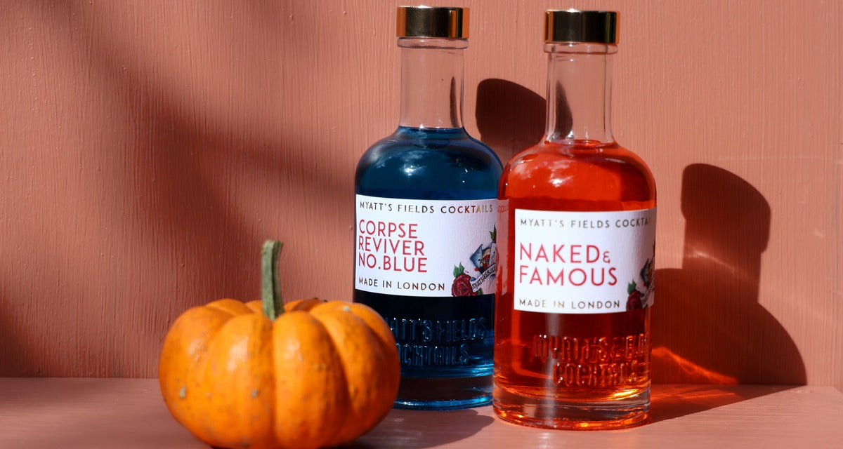 Two Myatt's Fields bottles, Corpse Reviver No. Blue and Naked & Famous, beside a small pumpkin against a terracotta wall in low autumn sun