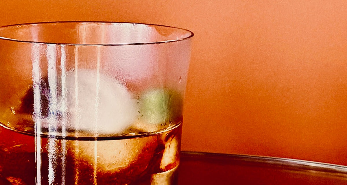 A Rum Old Fashioned in a frosted rocks glass with one large piece of ice and a strip of lime peel behind it