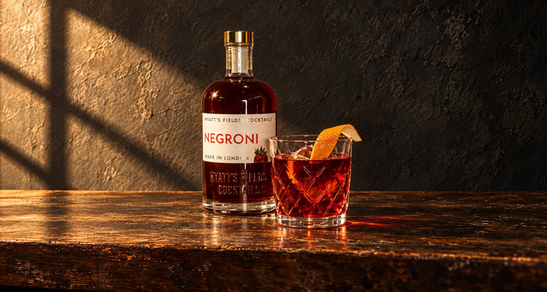 A bottle of the Myatt's Fields Negroni beside a Negroni over a large cube with a twist of orange peel, in low window light
